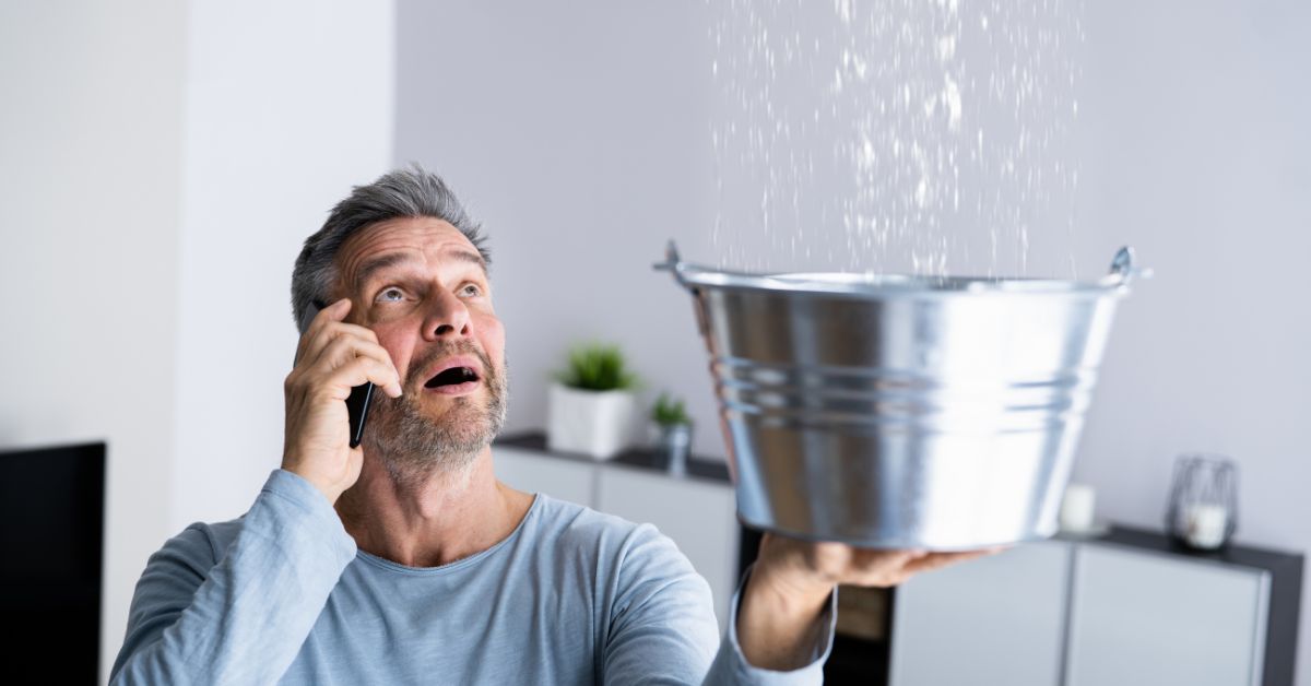 Man on his cellphone while holding up a bucket to catch water from a leaking ceiling